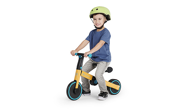 Ein kleiner Junge in blauem T-Shirt, Jeans und Sportschuhen sitzt auf einem gelben Kinderkraft-Dreirad. Er trägt einen Helm auf dem Kopf.