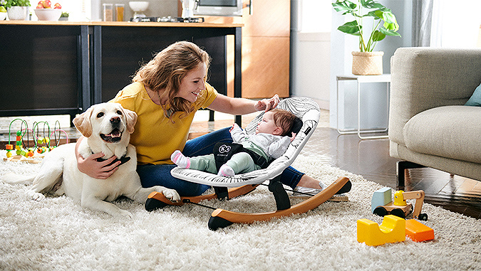 Die Mutter sitzt im Zimmer auf dem Boden und schaut auf das kleine Baby, das neben ihr in einer Kinderkraft-Liege liegt. Neben ihr liegt eine Spielmatte.