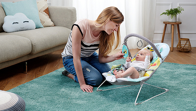 Die Mutter kniet auf dem Teppich und zeigt dem im Kinderkraftsitz liegenden Baby lächelnd das Spielzeug. Das Kind schaut mit Interesse zu.