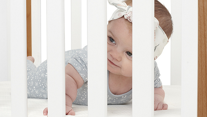 Ein mehrere Monate alter Säugling, der auf einer bequemen Matratze liegt, schaut durch die weißen Sprossen des Kinderkraft-Bettes.