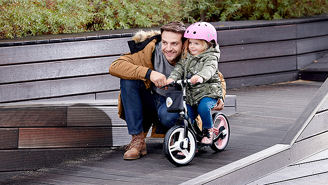 Ein Mädchen mit einem rosa Helm sitzt auf einem Kinderkraft-Dreirad. Sie wird von ihrem Papa am Sattel unterstützt. Sie schauen beide in die gleiche Richtung.
