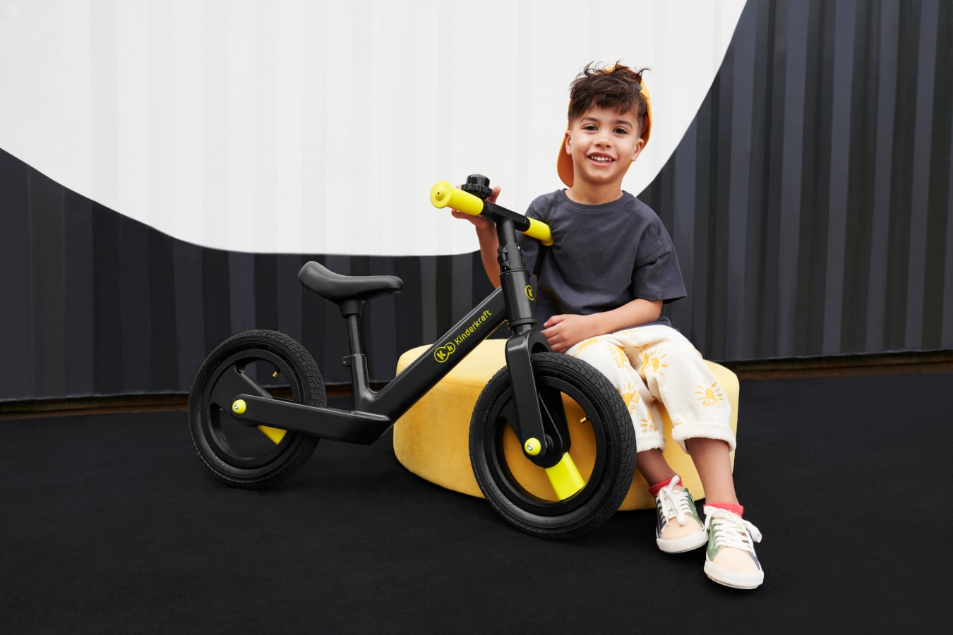 Ein entspanntes Kind sitzt in einem abstrakten Raum auf einem Sitzpuff und hält ein schwarzes zweirädriges Laufrad