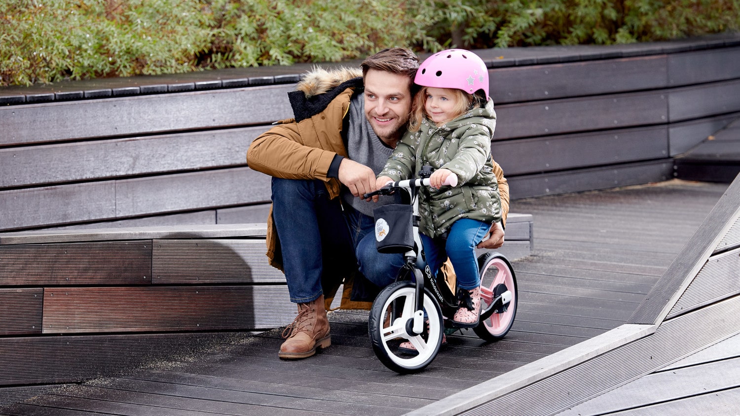 Ein Vater hockt im Herbst draußen neben einem Mädchen, das in einem Fahrradhelm auf einem zweirädrigen Laufrad in Schwarz und Rosa sitzt