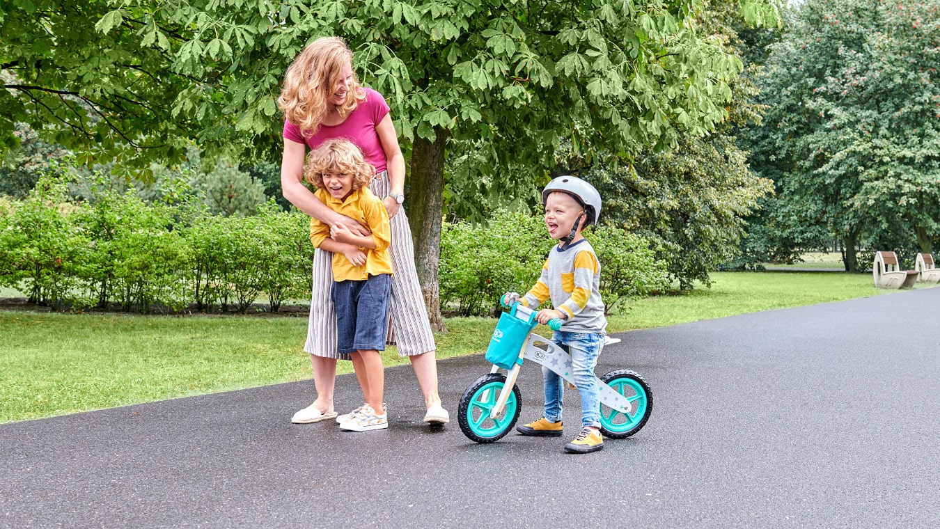 Eine Mutter geht mit einem Kind auf einem türkisfarbenen, zweirädrigen Laufrad und einem älteren Kind spazieren 