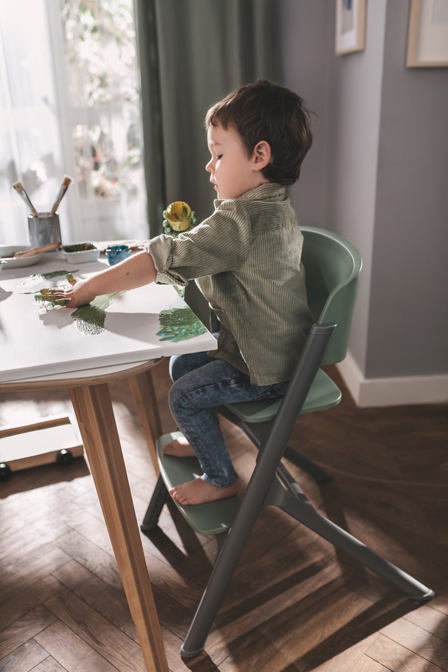 Ein Junge sitzt auf dem LIVY-Kinderhochstuhl von Kinderkraft, bemalt mit den Fingern  eine Leinwand, ist konzentriert und entspannt